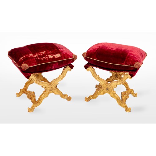 A pair of Italian giltwood carved folding stools, with a rectangular loose cushion seat, in a red silk velvet, above ‘X'-frame baluster supports, carved with scrolls, centred by a patera, joined by shaped stretchers centred by a stylised scallop-shell, on outward scrolled feet.